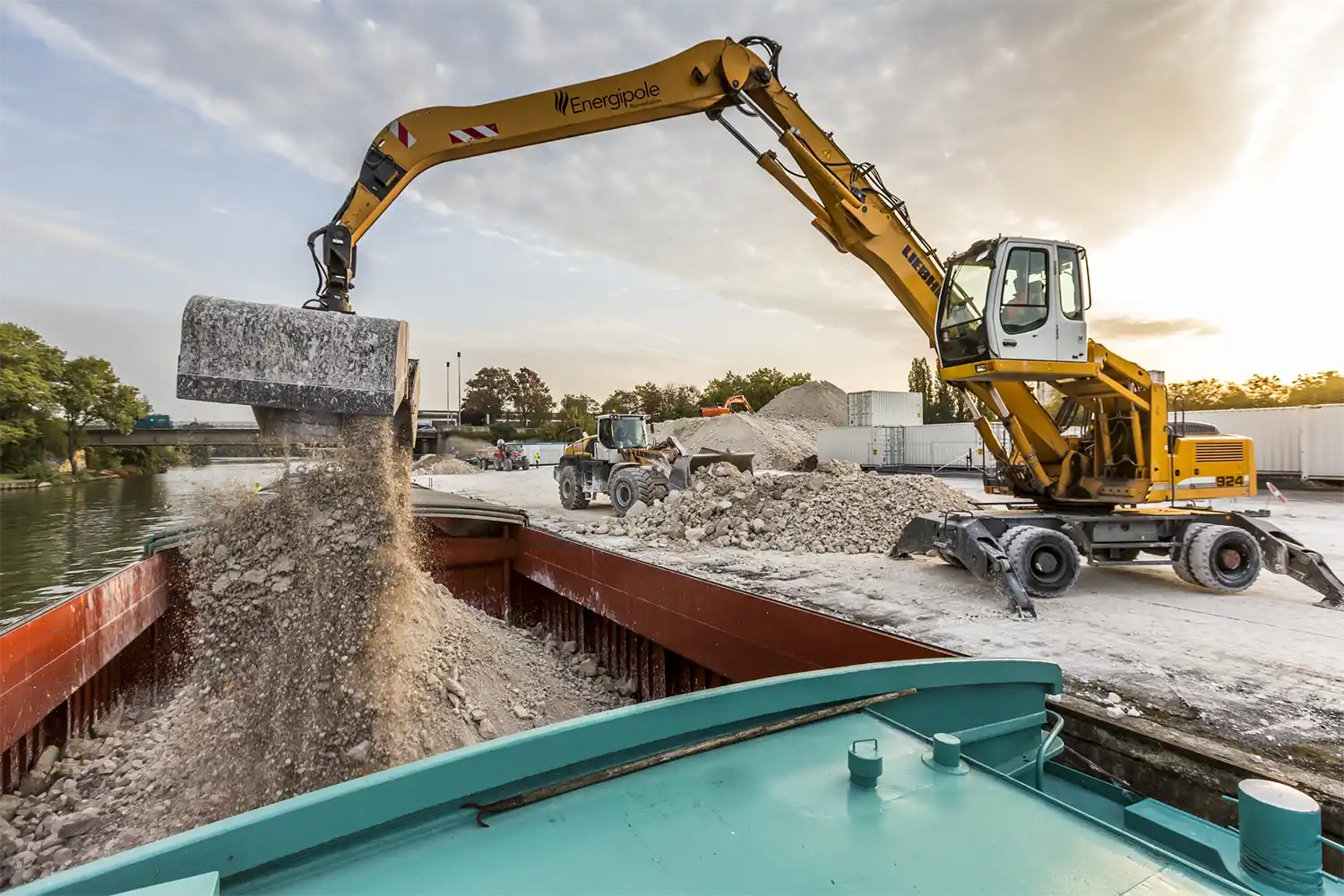 deblais-Plateformes-EppevilleOK Excavatrice jaune déchargeant des déblais sur une péniche, avec des tas de gravats et des conteneurs en arrière-plan sur le chantier d'Eppeville.
