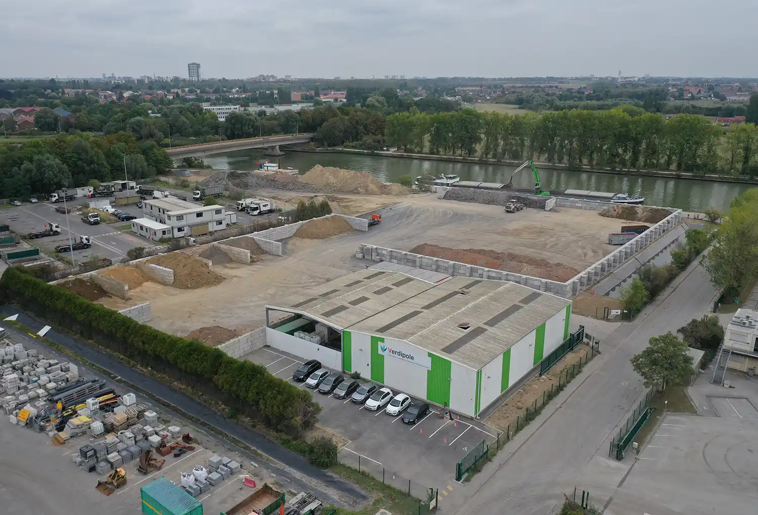 deblais-Santes Vue aérienne d'un site industriel avec un bâtiment blanc et vert, des voitures garées, des déblais et un canal bordé d'arbres.