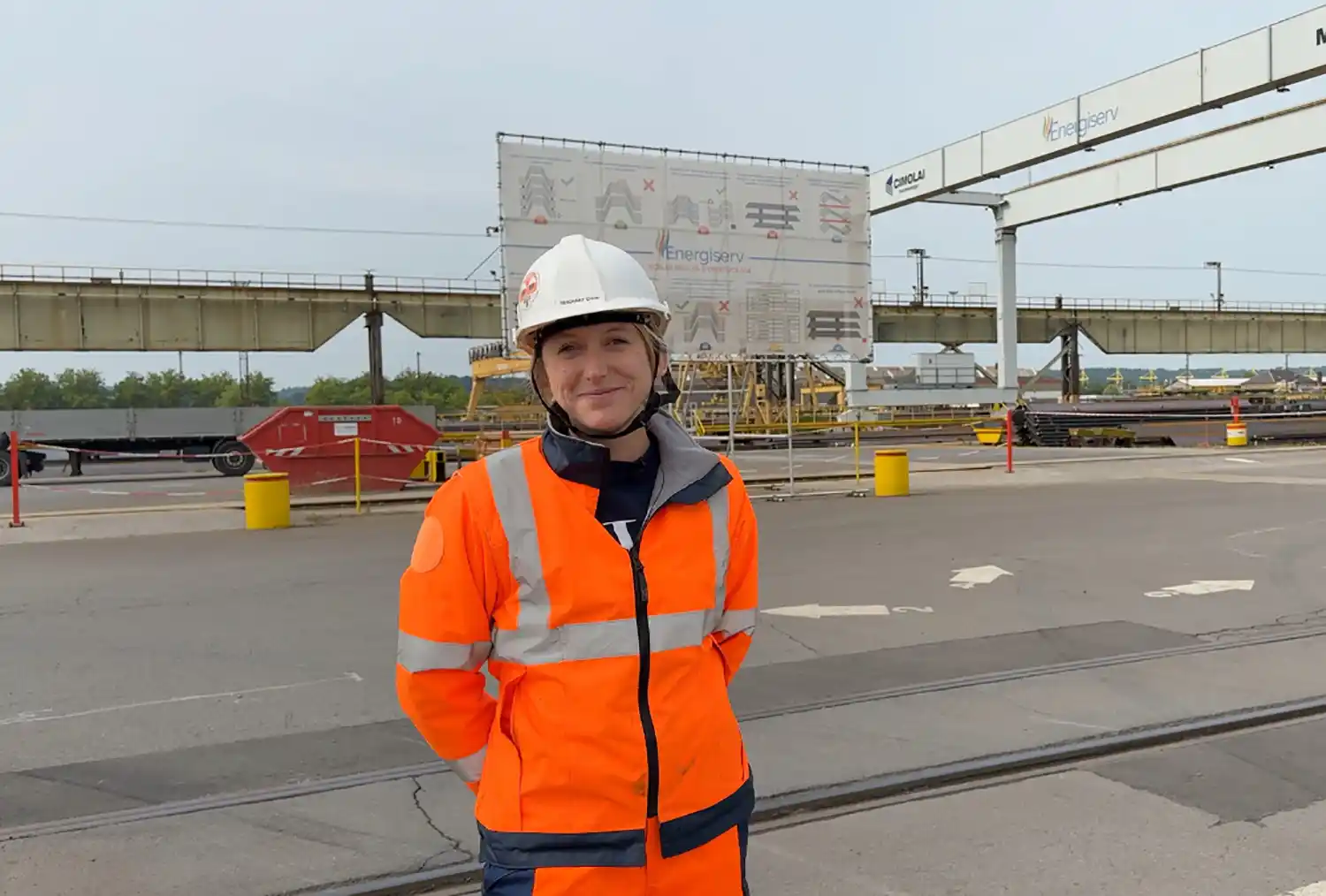 Personne en tenue de sécurité orange et casque blanc sur un site industriel avec structures métalliques et signalétique Energiserv en arrière-plan.