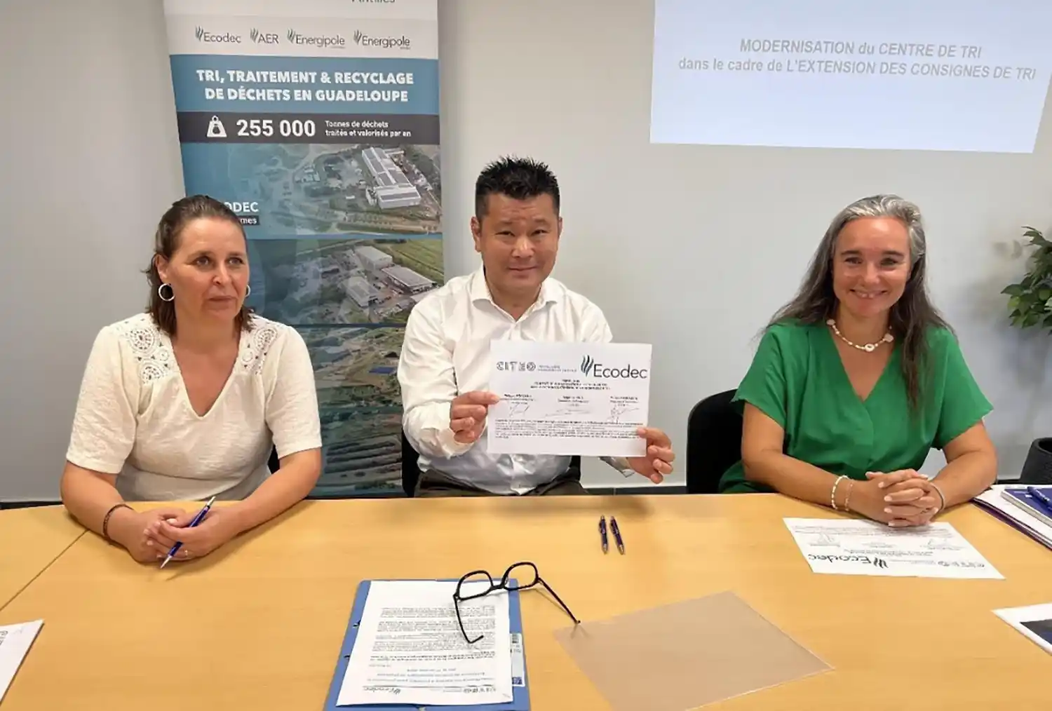 Trois personnes assises à une table, un homme tenant un document Ecodec, avec une affiche sur le tri des déchets en arrière-plan.