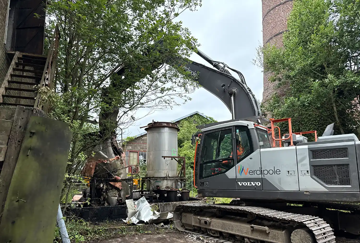 Une pelleteuse grise de marque Volvo avec le logo Verdipole soulève des débris près d'un bâtiment en brique et d'arbres.