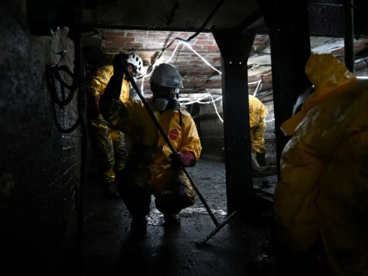 Des ouvriers en combinaison jaune et casque travaillent dans un espace sombre avec des murs de briques et des structures métalliques.