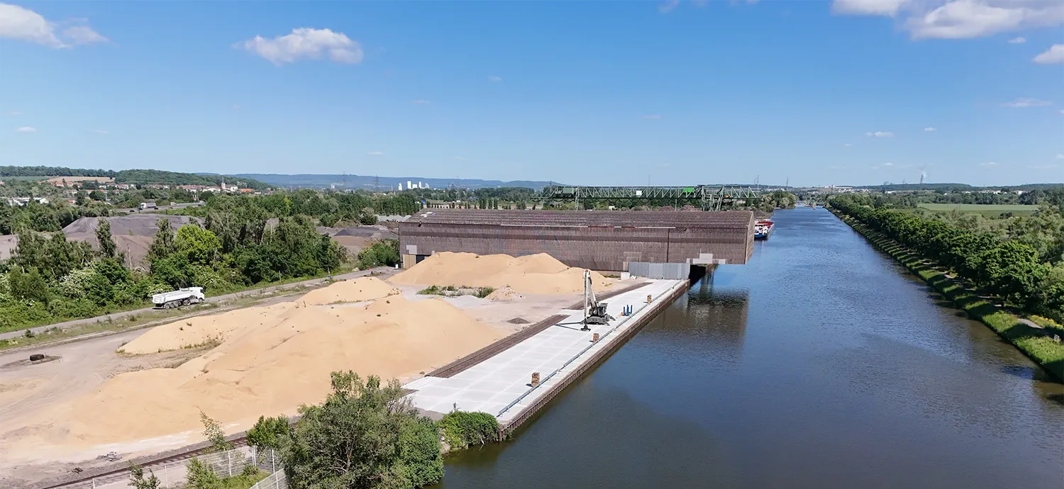 Site de stockage avec tas de sable, bâtiment industriel en arrière-plan et canal bordé d'arbres sous un ciel bleu.