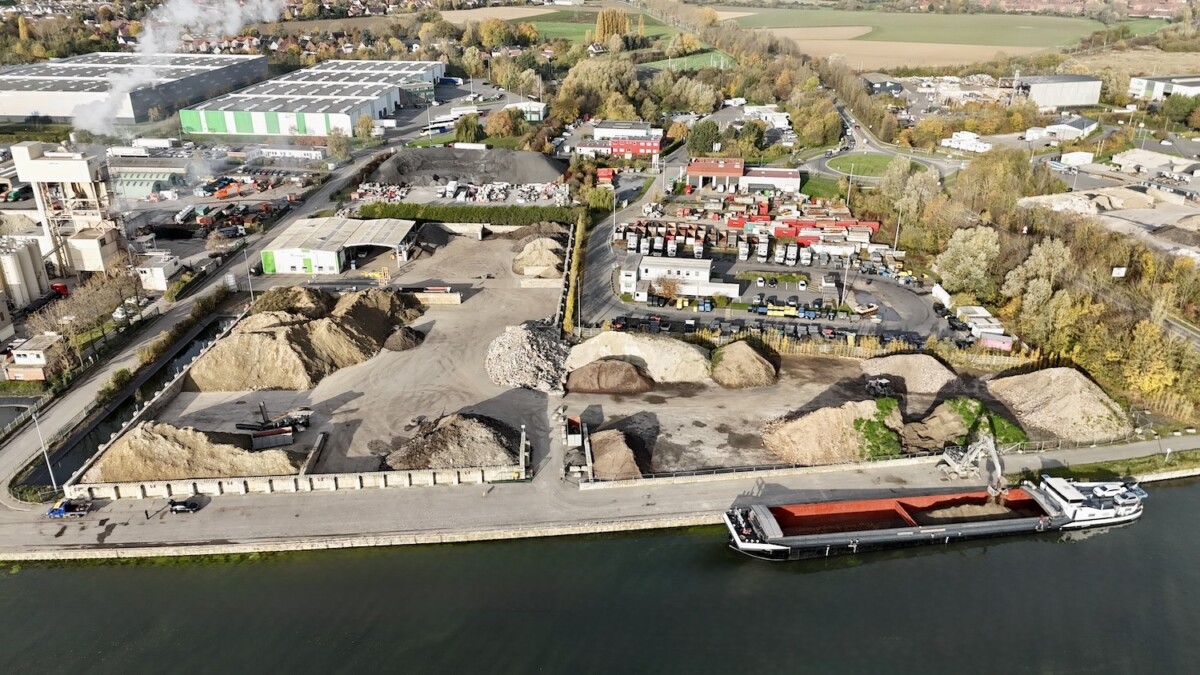 Vue aérienne d'un site industriel avec un bâtiment blanc et vert, des voitures garées, des déblais et un canal bordé d'arbres.